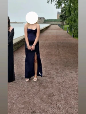 Marinblå långklänning med slits - Säljer min balklänning, endast använd 1 gång och inga täcken på användning. Snygg marinblå långklänning med smala axelband och öppen rygg med snörning. Klänningen har en hög slits framtill och en figurnära passform som framhäver siluetten. Perfekt för bal eller festliga tillfällen.