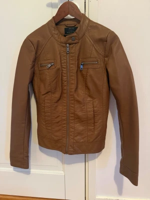 Brun skinnjacka från ONLY - Snygg brun ”fusk” skinnjacka från ONLY med två bröstfickor med dragkedja, ståkrage med tryckknapp och raka ärmar. Jackan har en klassisk bikerstil och dragkedja framtill. Perfekt för dig som vill ha en stilren och trendig look.