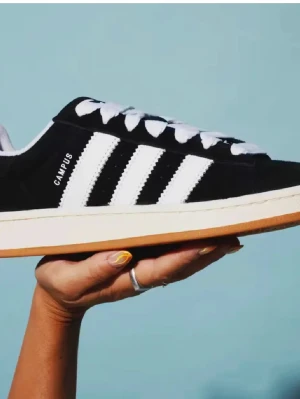 Adidas Campus svart/vit sneakers - Klassiska Adidas Campus sneakers i svart mocka med vita läderdetaljer och de ikoniska tre ränderna på sidan. Skon har vit sula med gummiyttersula och vita skosnören. Perfekt för dig som gillar retrostil och streetwear.
