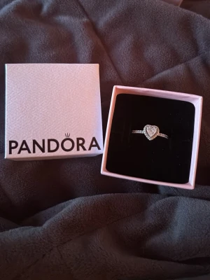 Silverring med hjärta från Pandora - Säljer en elegant silverring från Pandora med ett gnistrande hjärta i mitten och små stenar längs bandet. Ringen levereras i originalask och har en romantisk och stilren design som passar perfekt till dig som gillar klassiska smycken.