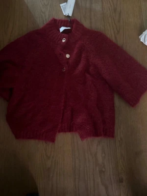 Röd kofta med knappar - Mysig röd kofta med tre knappar framtill och bred ribbad krage. Koftan har halvlånga ärmar och är gjord i ett fluffigt, stickat material som känns riktigt skönt. Perfekt för lager på lager och en chill look.