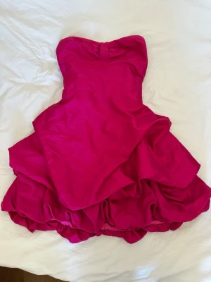 Axelbandslös rosa volangklänning - Säljer en axelbandslös kort klänning i stark rosa färg med volangdetaljer och knytband i midjan. Passar som en S