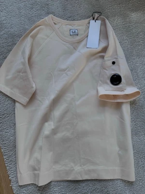 Beige t-shirt från C.P. Company - Säljer en beige t-shirt från C.P. Company med klassisk rund hals och korta ärmar. På ena ärmen finns en ficka med den ikoniska svarta linsen. Tillverkad i mjuk bomull för skön känsla och stilren look.