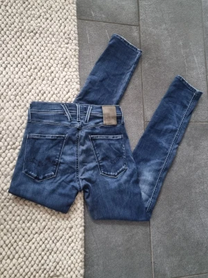 Replay Anbass - Snygga blå slim fit jeans från Replay med klassisk femficksdesign och slitna detaljer. Jeansen har normalhög midja, smala ben och är tillverkade i mjukt denimtyg med stretch för extra komfort. Perfekta för en avslappnad och trendig look.