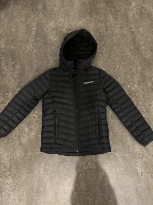 Svart dunjacka Peak Performance - Snygg svart dunjacka från Peak Performance med luva och dragkedja framtill. Jackan har quiltad design och diskret logga på bröstet. Perfekt för kalla dagar och enkel att matcha med olika outfits. Materialet är lätt och smidigt, vilket gör den bekväm att bära. Dunjacka är på bra skick och har inga synliga fläckar eller skador. Jag har använt den 1-2 säsonger och kedjan har förlorat lite färg men annars är den på mycket bra skick och inga problem med den. Den är tunn och bra för året om. 