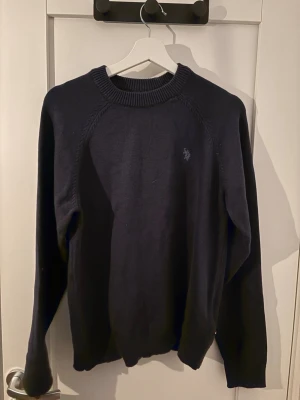 Mörkblå stickad tröja från U.S. Polo Assn. - Mörkblå stickad tröja från U.S. Polo Assn. med rund halsringning och diskret logga broderad på bröstet. Tröjan har långa ärmar och ribbade muddar vid hals, ärmslut och nederkant. Perfekt för lager på lager och enkel att matcha. Storlek S men passar M också