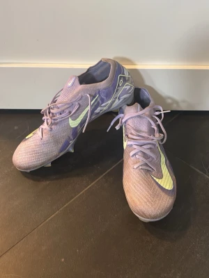 Lila Nike Mercurial fotbollsskor - Säljer ett par lila Nike Mercurial fotbollsskor med limegröna detaljer och snörning. Skorna har en låg profil och är tillverkade i ett lätt syntetmaterial för bra bollkontroll och grepp på planen. Perfekta för dig som vill sticka ut på fotbollsplanen.
