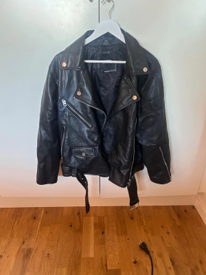 Svart biker skinnjacka med bälte - Snygg svart oversized skinnjacka från pull and bear med silvriga dragkedjor, tryckknappar och ett justerbart bälte i midjan. Säljer för att den tyvärr inte kommer till användning