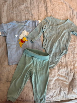 Sött babyset med Nalle Puh och body - Ett gulligt set för baby med en ljusblå t-shirt med Nalle Puh-tryck, en långärmad ljusblå body med små prickar och ett par matchande mjuka byxor. Perfekt för mysiga dagar hemma. Materialet känns mjukt och bekvämt mot huden.