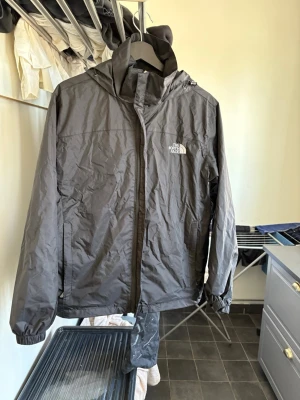Svart vindjacka från The North Face - Snygg svart vindjacka från The North Face med huva och dragkedja framtill. Jackan har elastiska muddar vid ärmsluten och logga på bröstet. Materialet är tunt och vindtätt, perfekt för blåsiga dagar. Innerficka med patch och justerbar nederkant.