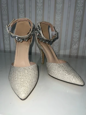 Glittriga pumps med silverdetaljer - Säljer ett par eleganta pumps i ljusbeige med glittrig yta och spetsig tå. Skorna har en smal klack och ett brett ankelband dekorerat med silvriga blommor och spänne. Perfekta för dig som vill sticka ut med stilrena detaljer.