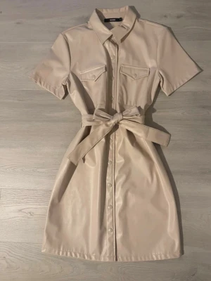 Beige skjortklänning i fuskläder S - Stilren beige skjortklänning från bikbok i fuskläder. Klänningen har korta ärmar, knappar framtill, två bröstfickor med lock och ett brett knytband i midjan för snygg siluett. Perfekt för dig som gillar trendiga och minimalistiska plagg.