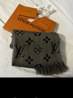 Grå halsduk från Louis Vuitton - Snygg grå halsduk från Louis Vuitton med klassiskt monogrammönster i svart. Halsduken har fransar i kanterna och är stickad i ett tjockare material som känns lyxigt och trendigt. Perfekt accessoar för att lyfta din outfit.