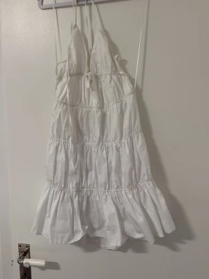 Vit kort sommarklänning med volang - Söt vit kortklänning med tunna axelband och volangkant nertill. Klänningen har omlottkänsla upptill och smockade sömmar som ger en luftig och avslappnad look. Perfekt för varma dagar och enkel att styla med accessoarer.