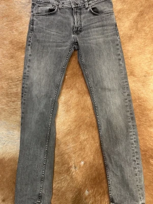 Grå nudie jeans - Snygga grå slim fit jeans med klassisk femficksdesign och diskreta slitningar. Jeansen har normal midja och raka ben, perfekta till en avslappnad streetstyle-look. Tillverkade i mjuk denim med stretch för extra komfort. W32, L32