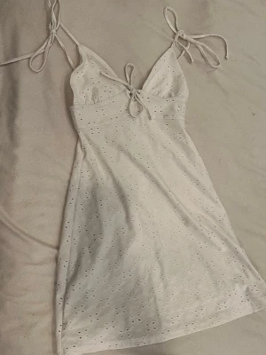 Vit spetsklänning med knytband - Söt vit klänning med broderad spets och små hålmönster. Klänningen har tunna axelband som knyts och en liten knytning framtill vid bysten. Perfekt för sommarens varma dagar. Och väldigt fin på midsommar.