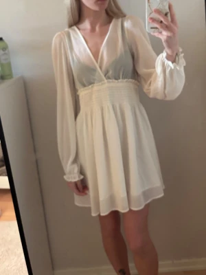 Vit omlottklänning med lång ärm - Söt vit kortklänning med omlott framtill och markerad midja. Klänningen har långa, transparenta ärmar med resår vid handlederna och ett lätt, flödande tyg som ger en romantisk känsla. Perfekt för sommarens alla tillfällen. (Finns exakt likadan i ljusblå längre ner i profilen)💕