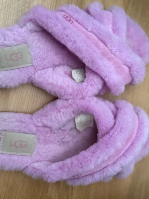 Lila fluffiga tofflor från UGG - Supermjuka och fluffiga tofflor från UGG i en härlig lila färg. Tofflorna har korsade band över foten och är tillverkade i ett gosigt syntetmaterial som känns lyxigt och varmt. Perfekta för att mysa runt hemma.