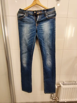 Nudie Jeans Tube Kelly - W28 L34 - Snygga blå jeans från Nudie Jeans Co med klassisk femficksmodell och kontrastsömmar. Jeansen har en smal passform och är tillverkade i slitstark denim med snygga slitningar och tvättade detaljer. Perfekta för en avslappnad och trendig look. Storlek W28 L34.