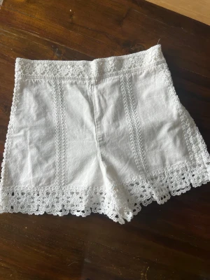 Vita spetsdetaljerade shorts - Säljer ett par vita shorts med fina spetsdetaljer längs kanterna och vertikala spetsband framtill. Shortsen har en hög midja och är tillverkade i ett lätt material som passar perfekt till varma dagar.
