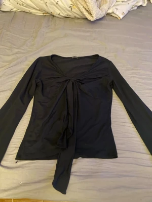Svart långärmad topp med knyt - Trendig svart långärmad topp med v-ringning och knytband framtill. Tunn och mjuk i materialet, perfekt för att styla med jeans eller kjol. Snygg passform som framhäver midjan och ger en cool look.