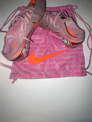 Rosa Nike Mercurial fotbollsskor - Säljer ett par rosa Nike Mercurial fotbollsskor med orange detaljer och svart logga. Skorna har snörning och är tillverkade i ett lätt syntetmaterial för bra bollkontroll. Kommer med en matchande rosa Nike-påse. Perfekta för dig som vill sticka ut på planen. 