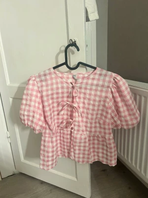 Rutig rosa blus med puffärm - Supersöt blus i rosa och vitt rutigt mönster med puffiga kortärmar och tre dekorativa knytband framtill. Aldrig använt! 