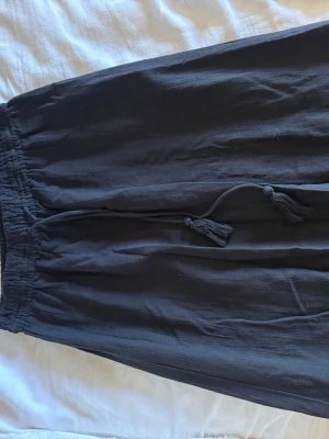 Svart långkjol med volang och snörning - Svart långkjol i mjuk bomull med resår i midjan och dekorativt knytband med tofsar. Kjolen har en bred volang nedtill.