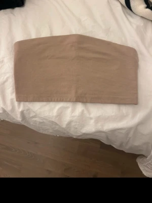 Beige bandeau tubtopp från 157 - Säljer en enkel och stilren beige bandeau tubtopp från 157. Toppen är axelbandslös och har en mjuk, stretchig känsla i bomullsmix. Perfekt att matcha med höga jeans eller kjol för en clean look.
