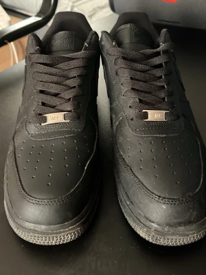 Nike Air Force 1 helsvarta sneakers - Säljer ett par klassiska Nike Air Force 1 i helsvart läder med matchande svarta skosnören och sula. Dom är använda mindre än 5 gånger. Ikonisk swoosh på sidan och metallbricka med 'AF1' på snörningen. Perfekta för dig som gillar stilrena och tidlösa sneakers.