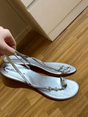 Silvriga kilklackssandaler från Matisse - Snygga silvriga sandaler från Matisse med kilklack och tunna remmar. Dekorativa kedjedetaljer framtill. Ovandelen är i glansigt material och sulan har en brun finish. 