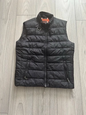 Svart dunväst från Superdry storlek S/M - Snygg svart dunväst från Superdry med quiltad design och hög krage. Västen har två dragkedjeförsedda fickor framtill och en cool detalj med spänne vid halsen. Västen är i ett väldigt bra skick. Tveka inte med att höra av dig vid fundering!