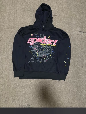 Svart hoodie med spindelnäts- och stjärntryck - Cool svart hoodie från Spider med färgglatt spindelnätsmotiv och texten 'spider!' framtill. Hoodien har tryckta stjärnor i gult, rosa och blått både fram och bak samt på ärmarna. Mjuk och bekväm med huva och ribbade muddar. Perfekt för dig som gillar streetwear och unika prints.