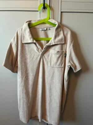Beige frotté sett från Warp - Säljer ett beige sett från Warp i mjuk frotté med krage och bröstficka. Perfekt för dig som gillar avslappnad stil och vill ha något annorlunda än vanliga t-shirts. Skön och enkel att matcha med det mesta.