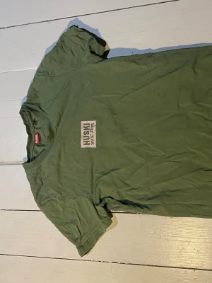 Grön t-shirt från Huski Wear - Säljer en grön t-shirt från Huski Wear med tryckt logga på bröstet. Klassisk rund hals och korta ärmar. Tillverkad i mjuk bomull som känns skön mot huden. Perfekt till jeans eller shorts för en avslappnad look.