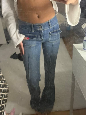 Blå bootcut jeans med fickdetaljer - Säljer ett par blå bootcut jeans med dubbla knappar och stora fickor framtill. Jeansen har markerade sömmar och en låg midja som ger en trendig look. Perfekta för dig som gillar en retro vibe och vill sticka ut lite extra.