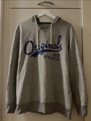 Grå huvtröja / hoodie från Jack & Jones - Storlek L - Snygg och bekväm grå hoodie från Jack & Jones säljes. Perfekt till vardags! Modell: JJORBROKE SWEAT HOOD CAMP. Storlek: L. Färg: Grå med vita dragsnören. Tryck: Blått tryck framtill – “Originals 1975 Athletics”. Material: 65% Polyester och 35% Bomull. Långärmad. Säljs i begagnat skick! OBS! En amatörlagning finns på ena ärmen (se bilder för att avgöra och bedöma dess skick!) I övrigt fullt användbar och skön!  Kika gärna in på mina andra annonser, jag säljer flera klädesplagg just nu!