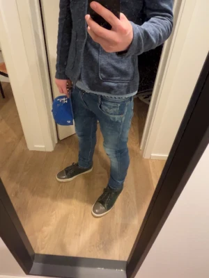 Blå slim fit jeans från Replay - Säljer ett par blå slim fit jeans från Replay med snygga slitningar och tvättade detaljer. Jeansen har klassisk femficksdesign, dragkedja och knapp i midjan samt Replay-logga på fickan. Perfekta för en avslappnad och trendig look.