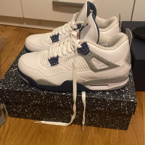 Nike Air Jordan 4 White Oreo - Nike Air Jordan 4 navy blue med vit ovandel i skinn, grå och marinblå detaljer samt röd Jumpman-logga på plösen och sulan. Klassisk siluett med snörning och meshpaneler på sidorna. Snygg kontrast mellan de olika färgerna och speckled midsole.
