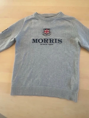 Grå tröja från Morris med logga - Snygg grå tröja från Morris med broderad logga och brittisk flagga på bröstet. Tröjan har rund halsringning, ribbade muddar och är tillverkad i mjukt material. Perfekt för en stilren och avslappnad look.