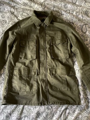 Olivgrön utilityjacka med dragkedja - Snygg olivgrön fieldjacket/overshirt med fyra stora fickor framtill, dragkedja och knappar. Jackan har en klassisk krage och är långärmad. Perfekt för dig som gillar en avslappnad men ändå stilren look. Hör av vid frågor och prisförslag 