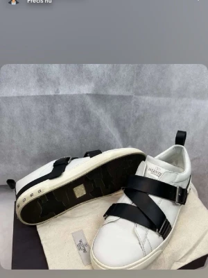 Valentino vita sneakers med svarta band - Snygga vita sneakers från Valentino med breda svarta läderband som korsar över foten och coola spännen på sidan. Skorna har en chunky vit sula och små nitar baktill. Perfekt för dig som vill sticka ut med en unik design.