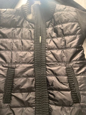 Cardigan  - Vadderad jacka | Svart | Med camouflagemönstrad insida Tuff svart vadderad jacka med ribbstickade detaljer längs kanterna och en cool camouflagemönstrad insida som syns när du öppnar den. Varm, bekväm och med en edge som skiljer den från mängden. Mer än möter ögat – kolla insidan! 🎖️