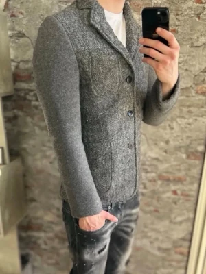 Grå ullkavaj med patchfickor - Stilren grå kavaj i ullmix med klassisk krage och tre knappar framtill. Kavajen har patchfickor och en bröstficka, samt mjuka, enfärgade ärmar i matchande grått. Perfekt för en dressad men avslappnad look.