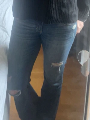 Lågmidjade hollister jeans med slitningar - Säljer ett par blå bootcut jeans med slitningar och hål på benen. Jeansen är utsydda av mig. Klassisk femficksdesign och låg midja. Perfekta för en avslappnad och trendig look. Materialet är denim i bomull. Har slitningar i skrevet men syns inte mycket när jeansen är på. Waist 29 & length 32❣️