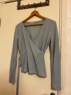 Ljusblå/Babyblå wrap top - Supersnygg ljusblå omlottopp med långa ärmar och ribbad struktur. Toppen har ett knytband i midjan som ger en smickrande passform och v-ringad halsringning. Perfekt att styla med jeans eller kjol för en trendig look. Använd en gång på Gröna Lund. Har en liten hål vid armhålorna, men går att sy om man vet hur (jag vet ej). 