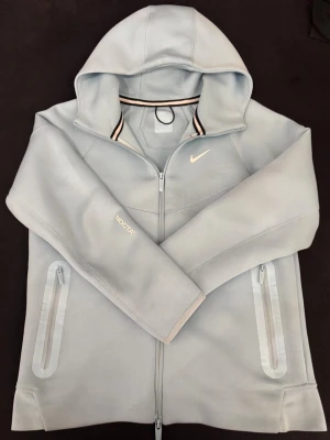 Ljusblå Nike NOCTA hoodie med dragkedja - Snygg ljusblå hoodie från Nike NOCTA med hel dragkedja, huva och två fickor med dragkedja. Diskret NOCTA-tryck på ärmen och klassisk Nike Swoosh på bröstet. Perfekt för en sportig och stilren look. Perfekt in för sommar den är i väldigt bra skick har inte använt den mycket.