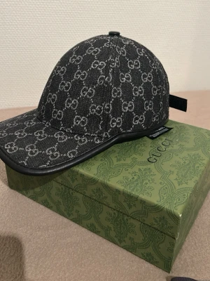 Ny keps från Gu*ci med monogram - Säljer en fin ny keps från Gucc* med det klassiska G*-monogrammet i grått över hela kepsen. Kepsen har böjd skärm, justerbart band bak och är tillverkad i ett slitstarkt tyg med läderdetaljer längs kanten. Perfekt för dig som vill ha en exklusiv och trendig accessoar.