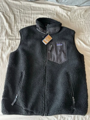 Svart fleeceväst från Patagonia - Säljer en svart fleeceväst från Patagonia med hög krage och dragkedja framtill. Västen har två sidofickor med dragkedja och en bröstficka i nylon med Patagonia-logga. Perfekt för lager-på-lager och riktigt skön att bära.