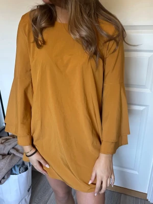 Kort senapsgul klänning med vida ärmar - Trendig kort klänning i senapsgult med vida, långa ärmar och rund halsringning. Klänningen har en lös passform och är tillverkad i ett mjukt bomullsliknande material. Perfekt för dig som gillar en avslappnad men stilren look.